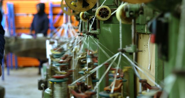 Close-up of threads running through textile machinery as a worker supervises the operation in an industrial factory. Suitable for content related to manufacturing, textile industry, factory operations, engineering, and production technology.