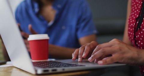 Close-up of colleagues working together in a modern office environment with a focus on teamwork and collaboration. A person is seen typing on a laptop while engaging in a conversation with a colleague. Ideal for use in articles or advertisements highlighting teamwork, casual businessings, or professional diversity in the workplace.