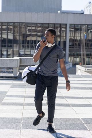 African American Young Man Walking in Athletic Wear Speaking into Smartphone on Plaza