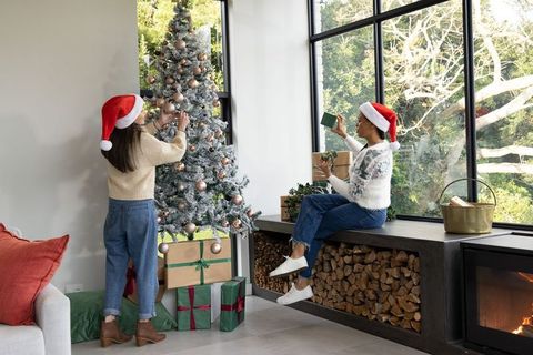 Female friends decorating snow-flocked christmas tree wearing santa hats wrapping gifts