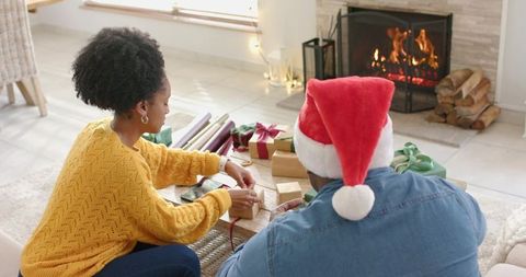 Couple wrapping holiday gifts by fireplace wearing santa hat, cozy fireside warm moment