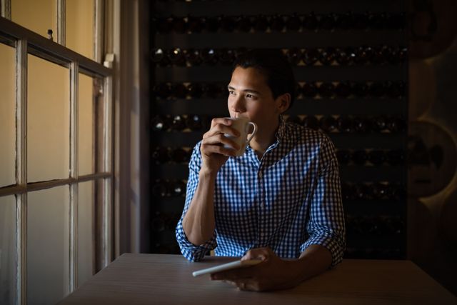 Man sitting by window in cozy restaurant, drinking coffee and holding tablet, appearing thoughtful. Ideal for use in lifestyle blogs, technology advertisements, relaxation and leisure promotions, or articles about modern dining experiences.