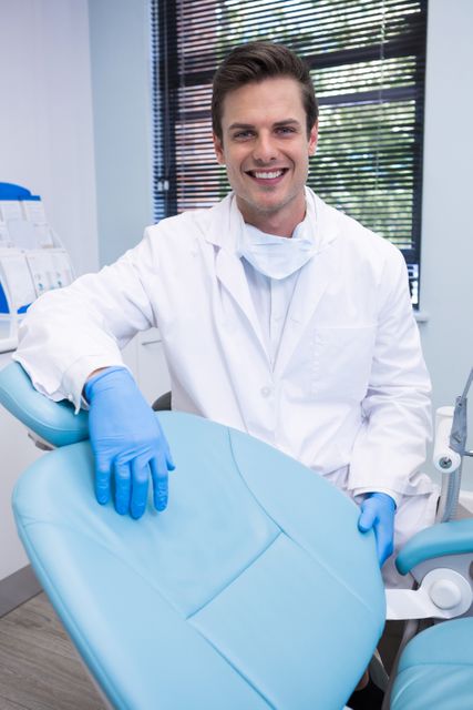 Dentist standing by a dental chair in a modern clinic, smiling confidently. Ideal for use in healthcare, dental care, and medical professional contexts. Can be used for promoting dental services, illustrating articles about oral health, or enhancing medical websites.