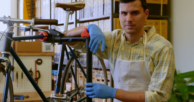 Man dressed in apron and blue gloves holding bicycle in workshop. Suitable for use in articles about bicycle repair, cycling maintenance, professional workshops, and small business promotions.