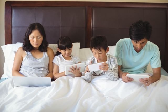 Family sitting in bed using various electronic devices including a laptop, tablet, and smartphone. The parents and children are engaged with their screens, showcasing a modern lifestyle. Ideal for use in articles about family life, technology use, digital age, and modern parenting.