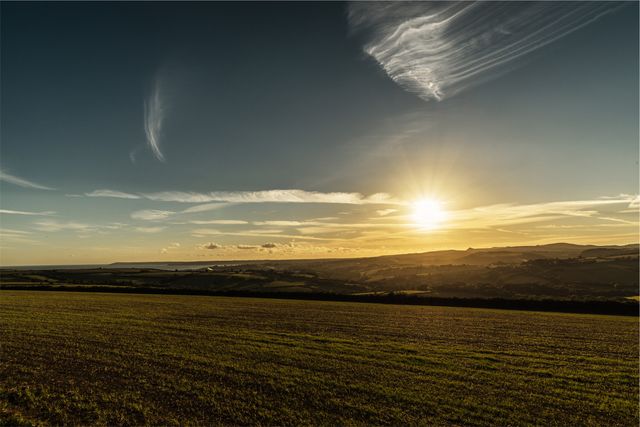 Wide-angle view of countryside during sunset, presenting a stunning horizon and expansive fields. Light clouds enhance the gradient sky from orange to deep blue, showcasing the beauty of nature. Ideal for use in background images, travel blogs, environmental presentations, or inspirational contexts promoting tranquility and serenity.