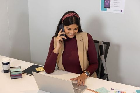 Asian professional multitasking, talking on smartphone while typing on laptop at desk