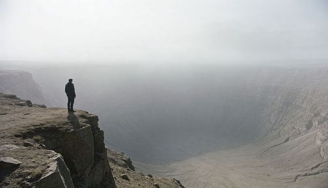 Solitary hiker standing on foggy cliff edge overlooking vast crater and desolate plateau