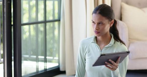 Contemplating woman holding tablet while seated in minimalist sunlit living room