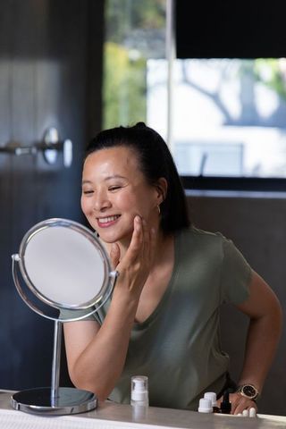 Asian woman applying skincare at vanity smiling while checking mirror in morning routine