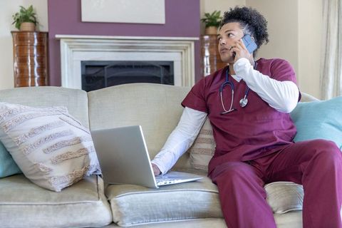 African american male nurse working remotely on sofa, talking on phone and using laptop