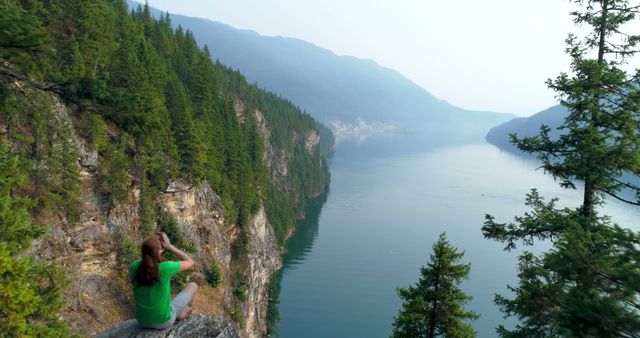 Woman sits on rocky clifftop, overlooking tranquil mountain lake and dense evergreen trees. Ideal for promoting outdoor adventures, hiking gear, travel destinations, nature retreats, or wellness retreats. Captures natural beauty and serene landscape, perfect for travel blogs, vacation brochures, and environmental campaigns.