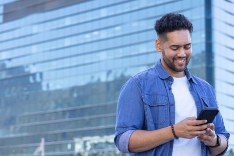 Young man smiling and checking smartphone outside modern glass office building