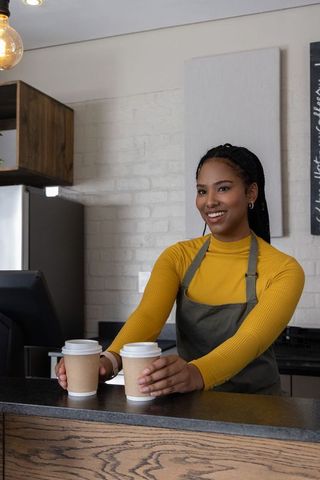 African american barista serving two to-go coffee cups while smiling at counter in cozy cafe