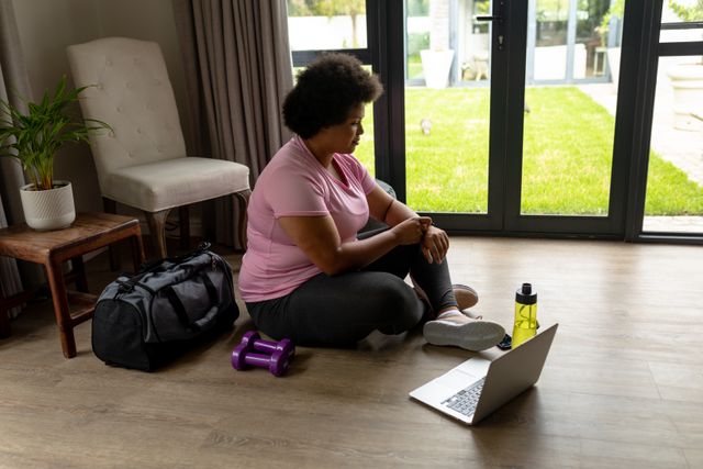 African American woman sitting on floor using laptop for home workout. She is wearing casual fitness attire and a wristwatch, with dumbbells and a water bottle nearby. Ideal for content on home fitness, online exercise programs, and healthy lifestyles.