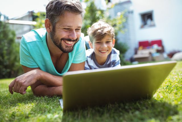 Father and son lying on grass in garden using laptop, both smiling and enjoying time together. Ideal for concepts of family bonding, outdoor activities, technology use, and leisure time. Suitable for advertisements, blogs, and articles related to parenting, family life, and summer activities.