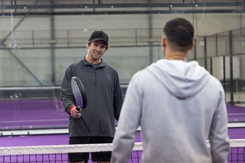Friendly indoor padel match on purple court with two players smiling and rallying near net
