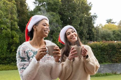 Two diverse women wearing santa hats holding steaming mugs, smiling outdoors, holiday sweaters
