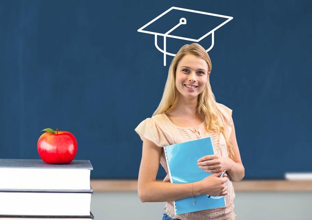 Happy teenage girl holding book with mortarboard head smiling at camera in the classroom