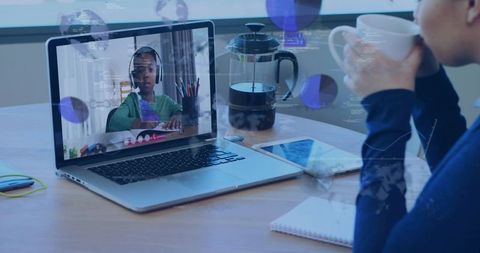 Parent watching child remote learning on laptop while sipping coffee at sunlit home desk