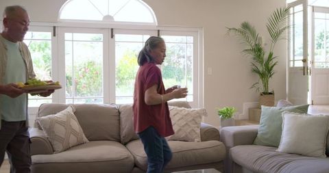 Senior couple carrying snacks and drinks into bright natural living room with plants