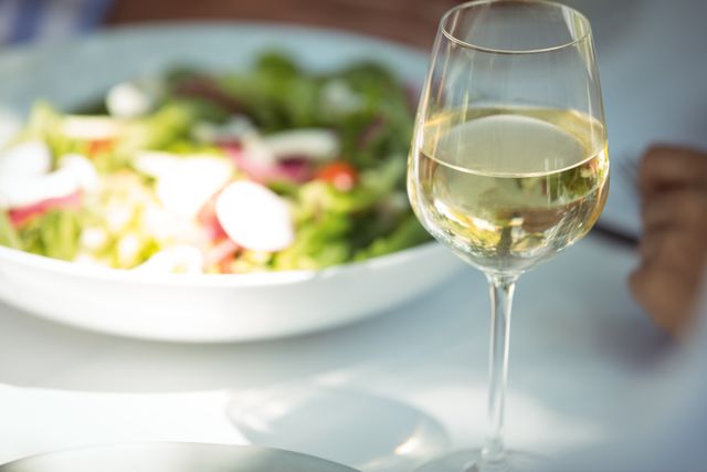 This image captures a close-up view of a wine glass filled with white wine and a fresh salad on a dining table in a restaurant. Ideal for use in articles or advertisements related to dining, gourmet food, healthy eating, restaurant promotions, or lifestyle blogs. It conveys a sense of elegance and culinary delight.