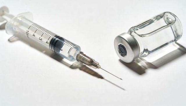Syringe and vaccine vial resting on white surface with uncapped needle and clear liquid