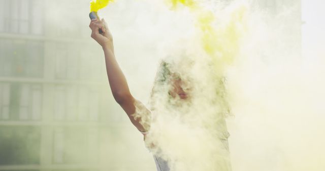 Person holds yellow smoke bomb, creating dense smoke cloud in urban area. Useful for illustrating themes such as celebration, activism, festivals, demonstrations, or urban art events. Could add dynamic and vibrant visuals to articles, promotional materials, or social media posts about urban culture and events.