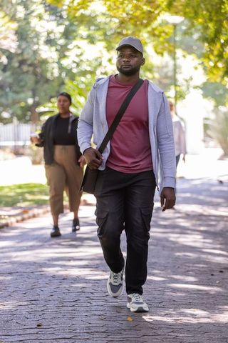 Urban commuter walking on shaded tree-lined path wearing cap and crossbody bag