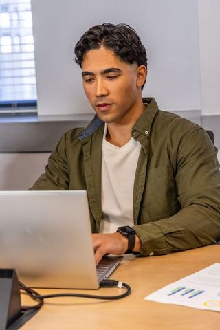 Focused asian man working on laptop at desk wearing olive overshirt in modern office