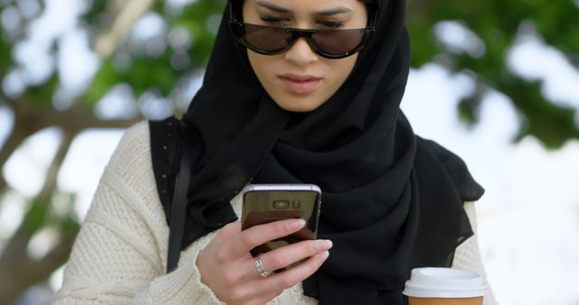 Muslim woman checks her phone outdoors, with copy space. She balances a coffee cup while focusing on her smartphone.