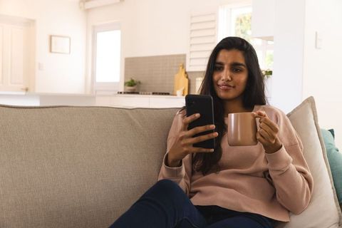 Relaxed Woman Enjoying Coffee While Using Smartphone on Sofa
