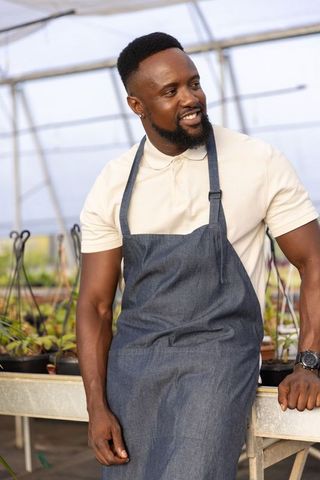 Smiling african american gardener leaning on bench in greenhouse wearing denim apron