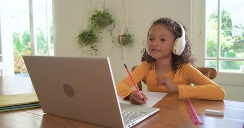 Girl studying online wearing headphones writing notes at laptop in bright room