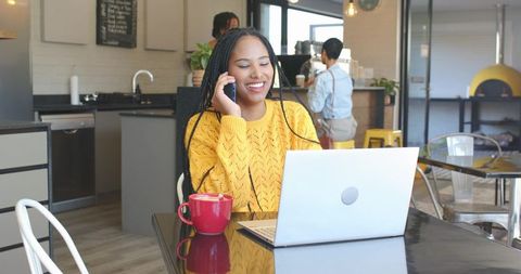 Smiling woman working remotely on laptop and phone in cozy modern cafe with red mug