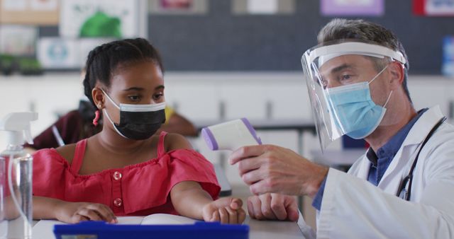 A doctor wearing a lab coat, face mask, and face shield measures the temperature of a girl, who is also wearing a mask, in a classroom. A hand sanitizer bottle is visible. Useful for illustrating safety precautions in schools, healthcare measures during the pandemic, or educational environment safety protocols.