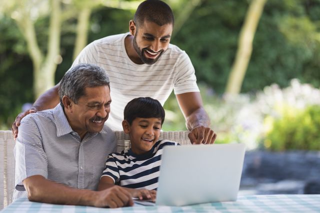 Three generations of a family enjoying time together outdoors, using a laptop on a porch. The grandfather, father, and son are smiling and bonding over technology. This image can be used for family-oriented content, technology in family life, intergenerational relationships, and leisure activities.
