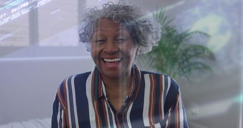 Smiling senior woman wearing striped shirt and necklace sitting on sofa in sunlit living room