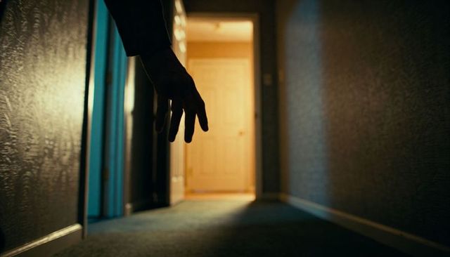 Shadowy hand reaching through dim corridor toward warm lit doorway cinematic suspense