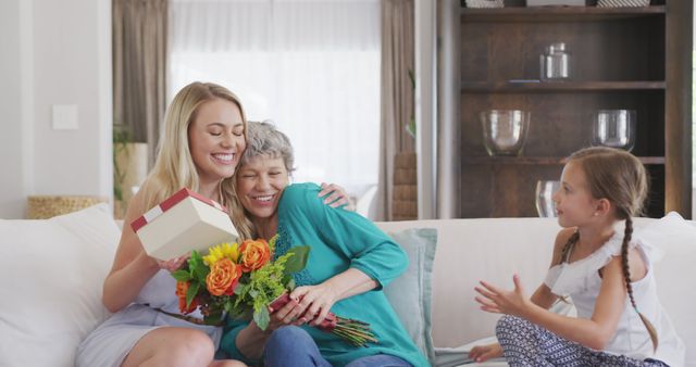 Happy caucasian grandmother, mother and daughter sitting on sofa, embracing and giving present. Mother's day, celebration, domestic life, family, and togetherness.