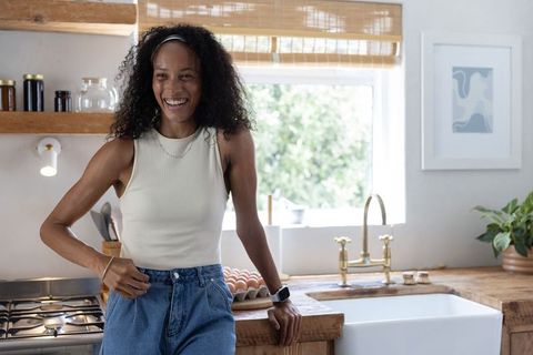 African american woman smiling and leaning on farmhouse sink holding egg carton in kitchen