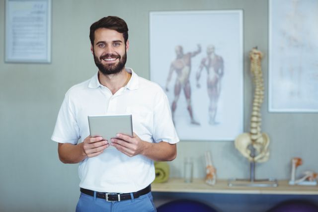 This image shows a smiling physiotherapist holding a digital tablet in a clinic. The background includes anatomy posters and a spine model, indicating a medical setting. This image can be used for healthcare websites, physical therapy promotions, medical blogs, and wellness articles.