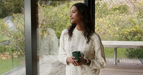 Cozy woman gazing through glass door holding mug on wooden deck overlooking lemon trees