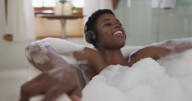 Man smiling while relaxing in a bubble bath, listening to music through headphones. This can be used for concepts of self-care, relaxation, and enjoyment, perfect for promoting spa services, personal care products, or mental health awareness. The setting offers a warm and inviting atmosphere.