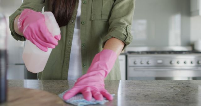 Woman cleaning kitchen counter with spray bottle and pink gloves, emphasizing hygiene, cleanliness, and home maintenance. Suitable to illustrate household chores, domestic services, and hygiene practices for magazines, blogs, or advertisements related to cleaning products and home care.