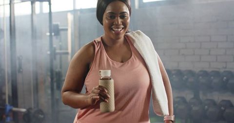 Smiling african american woman holding water bottle and towel after workout in gym
