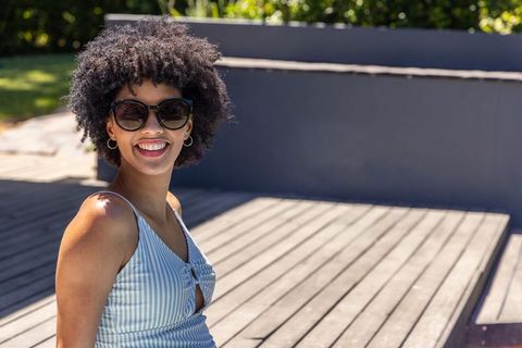 Smiling african american woman wearing sunglasses and striped dress on sunlit wooden deck