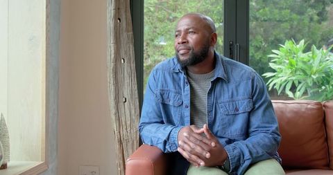 Middle-aged african american man gazing out window on leather sofa in sunlit living room