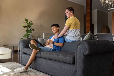 Smiling male couple relaxing on sofa sharing tablet and affectionate moment in sunlit home