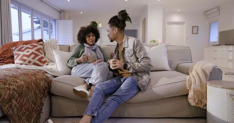 African american couple sitting on sofa holding mugs, chatting in cozy modern living room